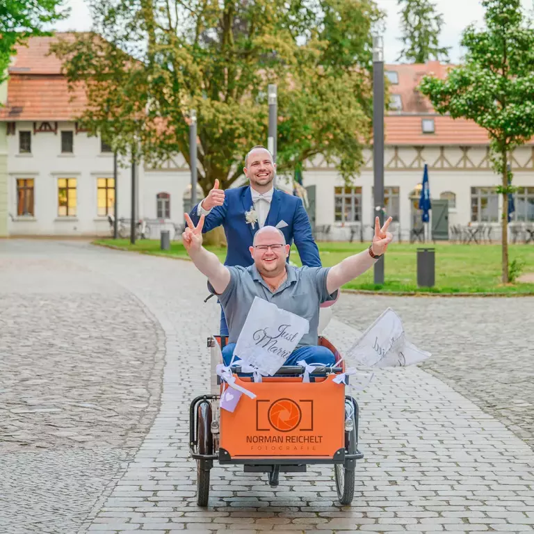 Bräutigam im Anzug in einem geschmückten Lastenrad mit 'Just Married'-Schild, geschoben von einer anderen Person, vor historischer Kulisse.